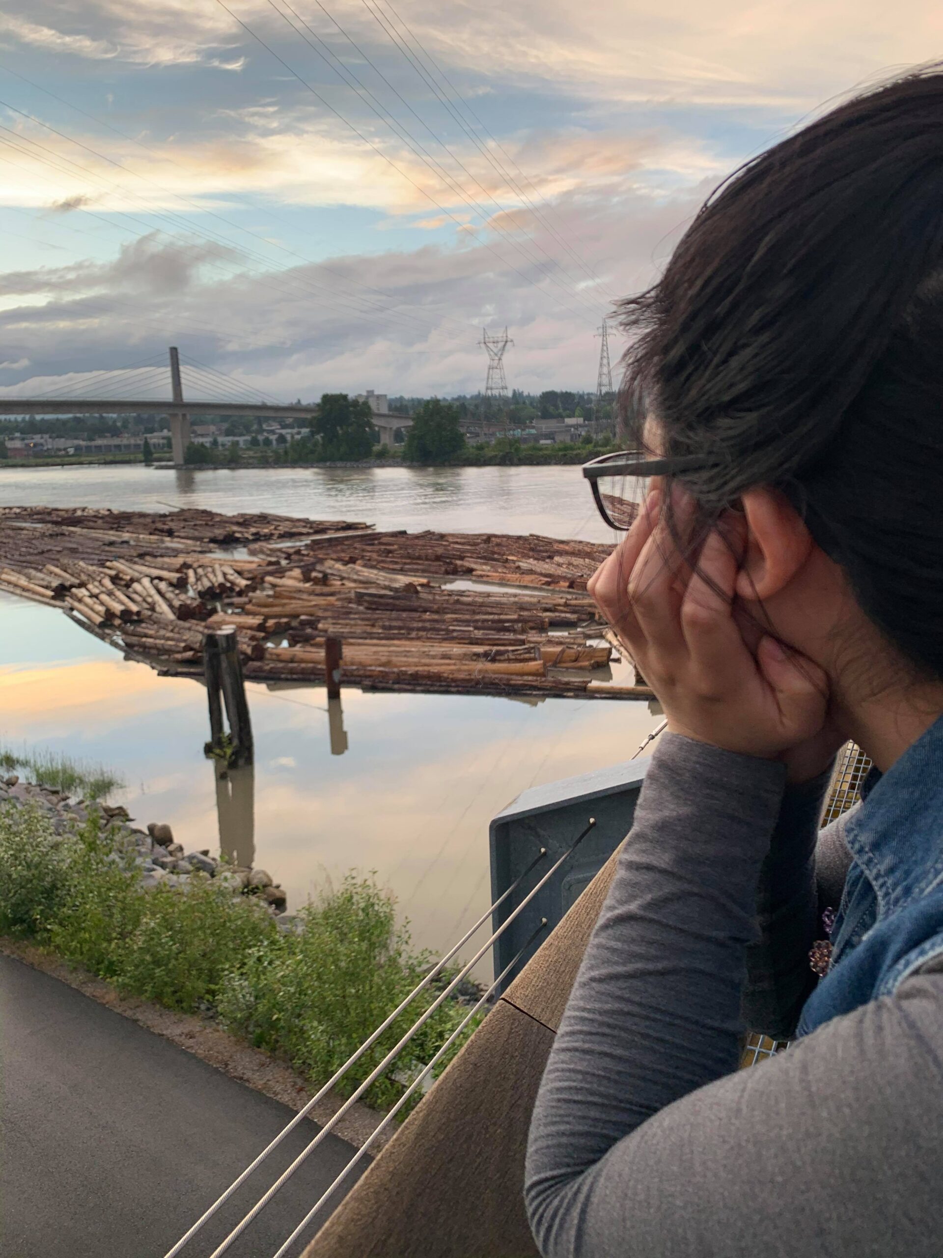 An Asian woman with glasses and her hair tied back in a grey long sleeve and blue denim vest looking into the sunset along a river. Her face, in profile, is resting in her left hand.